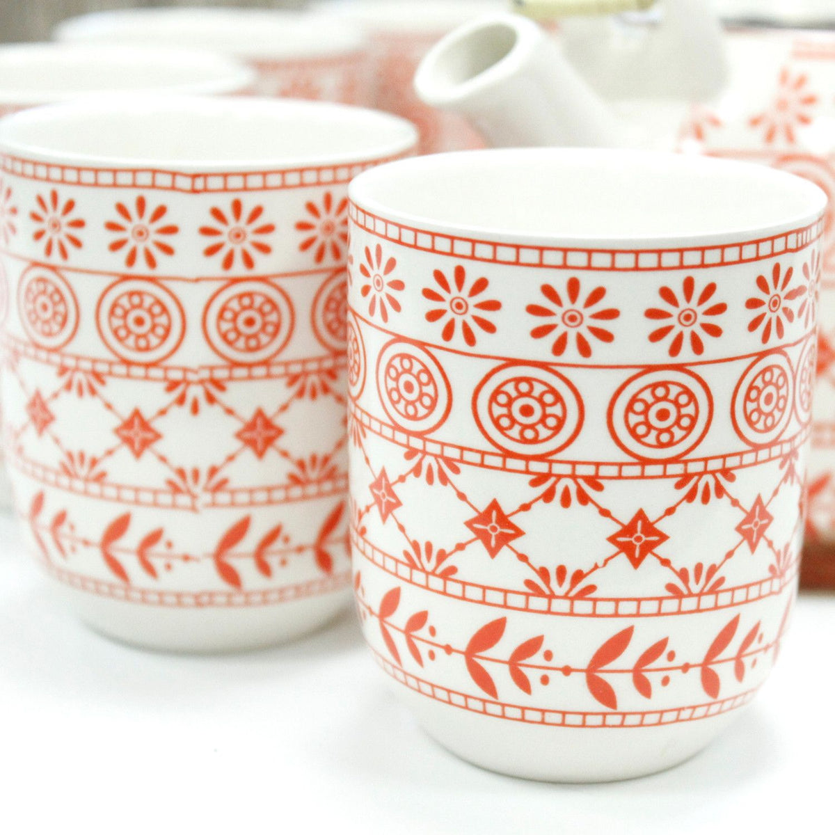 Teeservice-Set mit rötlichem Muster auf weißem Keramik, bestehend aus Teekanne und sechs Tassen mit dekorativen Motiven.